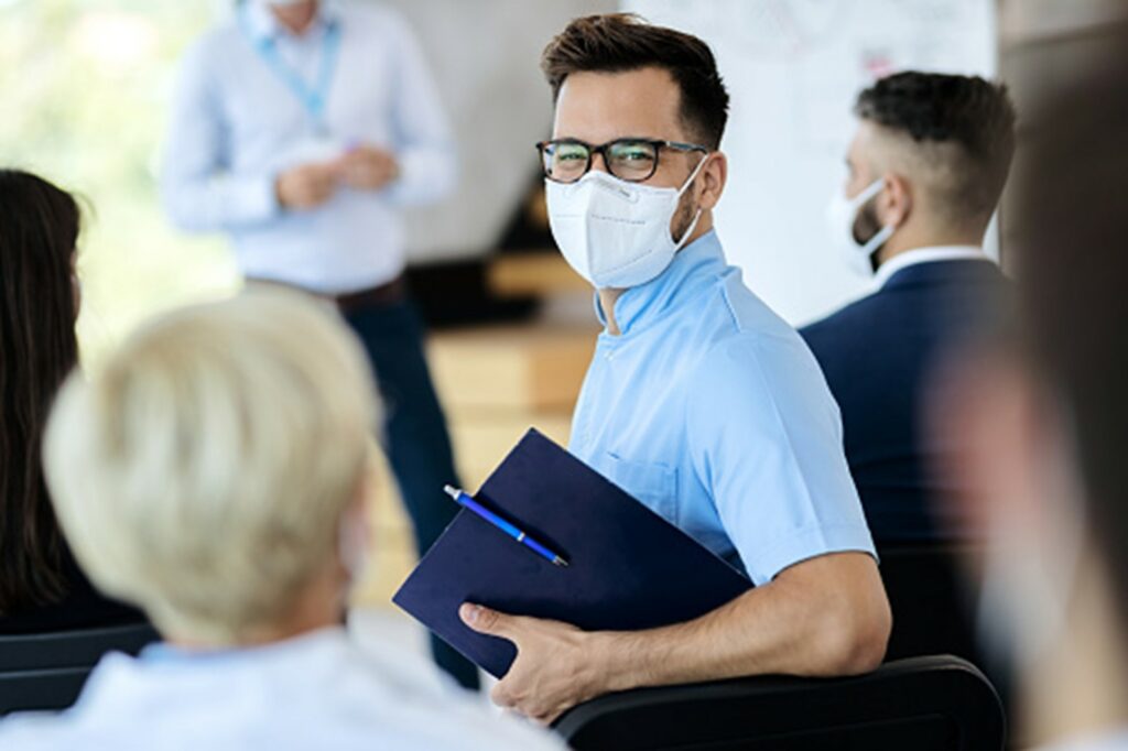 Ein Mann mit Mund-Nasen-Schutz und hellblauem Hemd steht im Fokus zwischen mehreren Menschen und hält eine Mappe in der Hand.